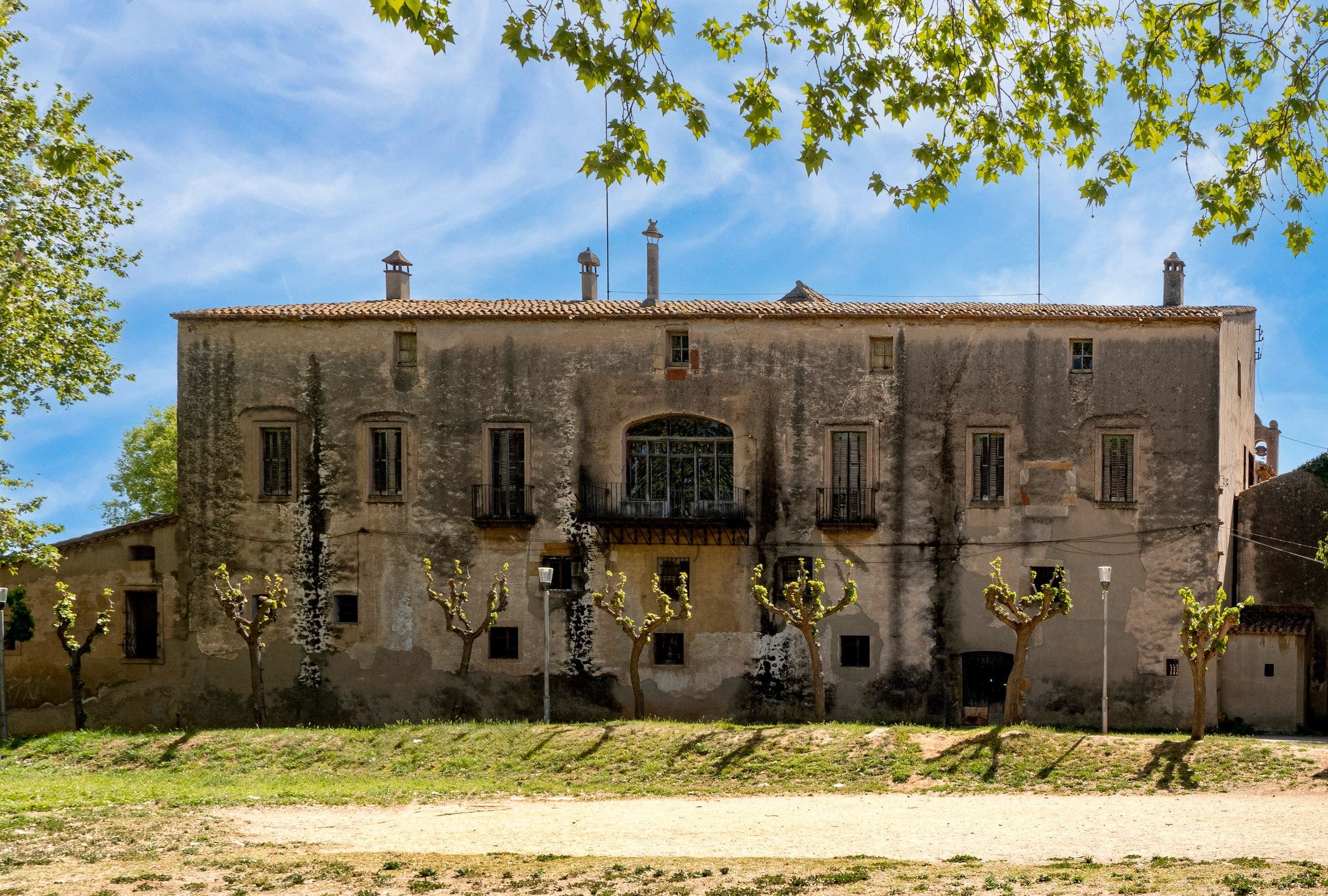 Façana d'una masia gran de pedra amb arbres al davant i cel blau